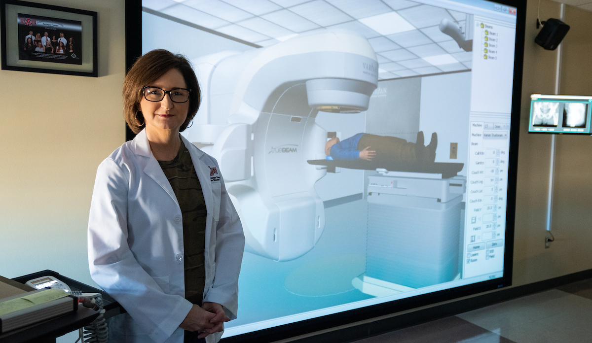 Instructor in white lab coat stands next to Virtual Environment Radiotherapy Training (VERT) system