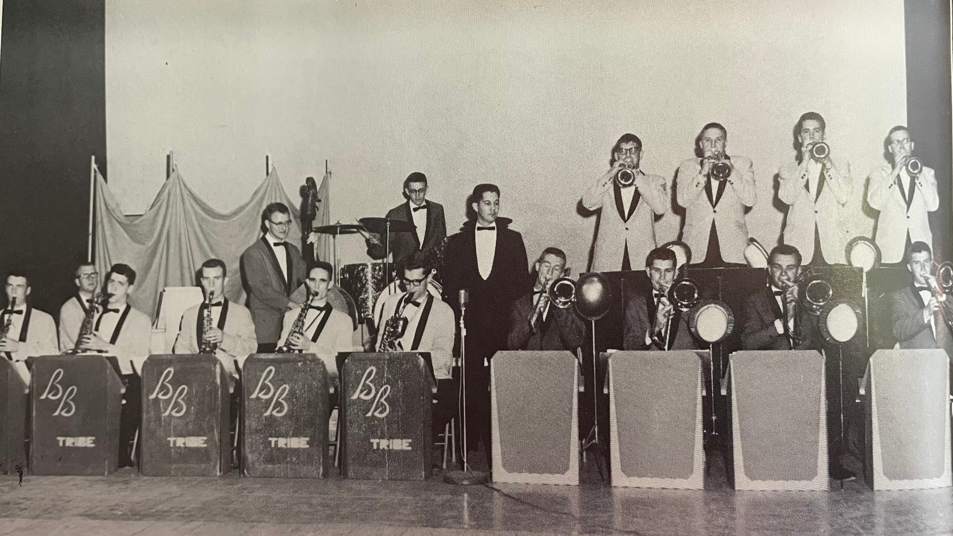 Historical photo of the A-State Jazz Band from the 1960s.  The front row contains saxophones and trombones seated behind box stands, the second row contains standing strumpets and the rhythm section.