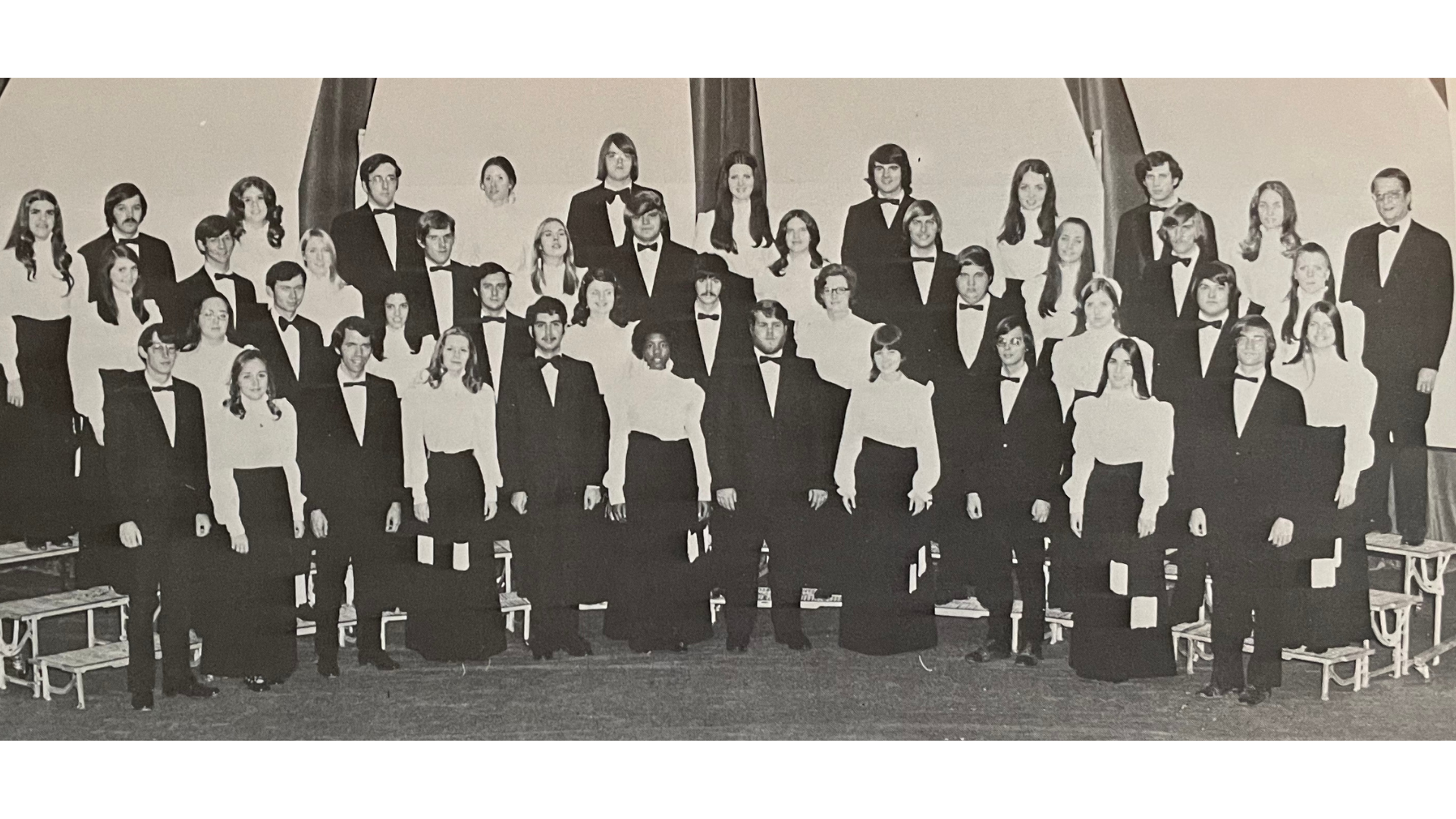 Historical photo of the A-State Concert Choir on risers.  Women are in white tops and black shirts, men are wearing tuxedos.