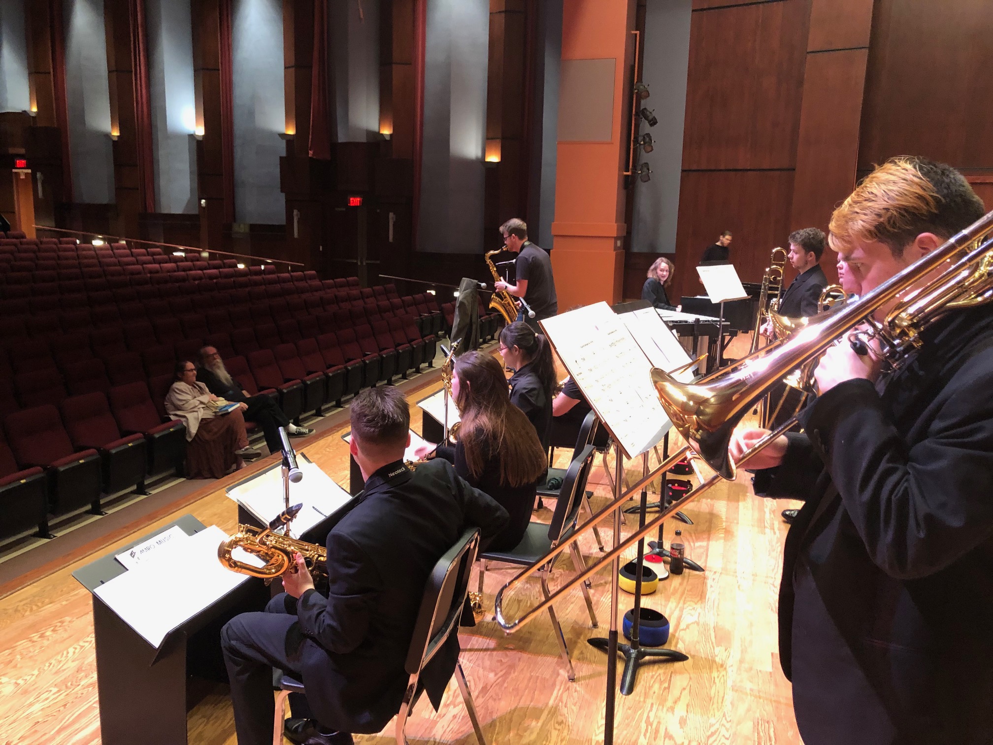 Side view of a jazz band on the Riceland Hall stage.  In the forefront is a trombone player and the line of saxophones.  A saxophone is at a microphone soloing. .