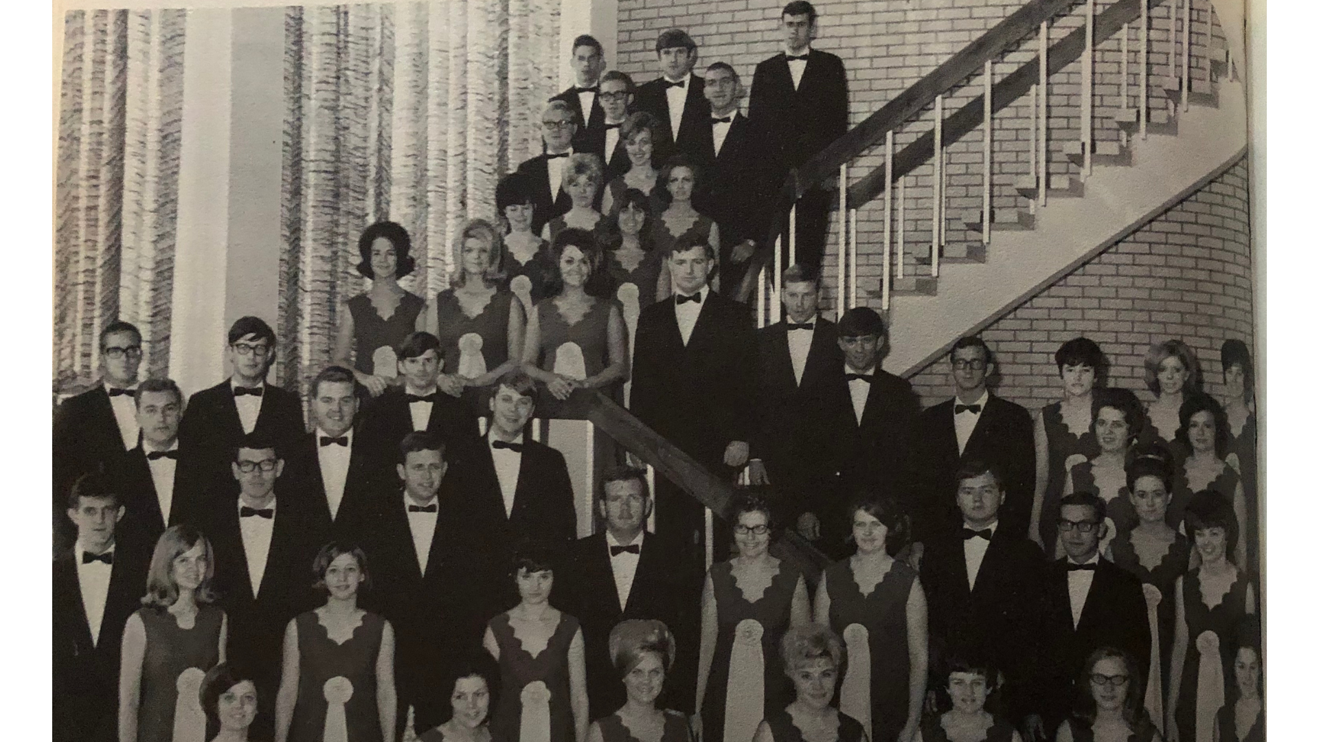 Historical photograph of an A-State choir around the grand staircase in the front of the Fine Arts Center.  Students are wearing their two toned dresses and tuxes.