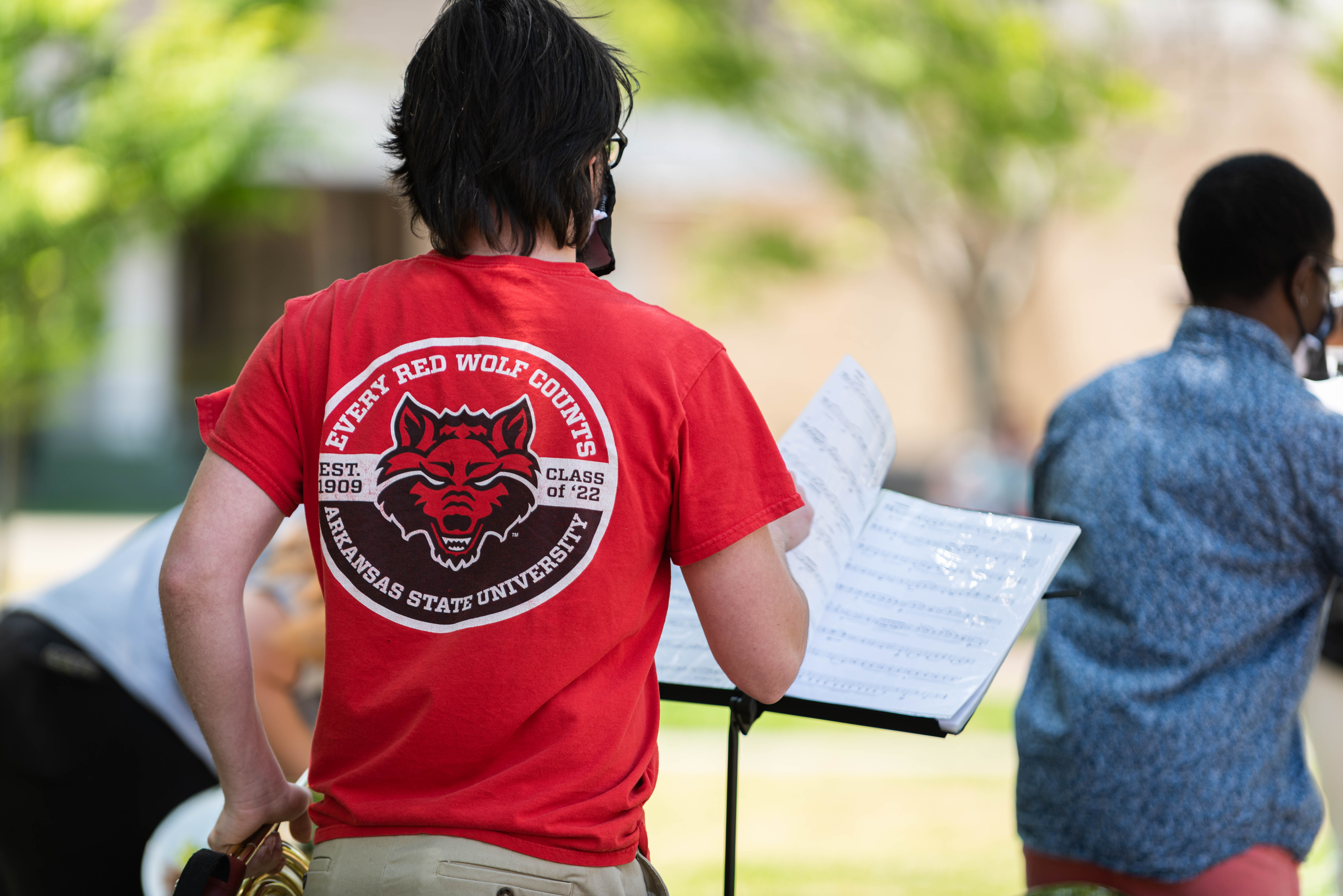 Looking at the backs of two french horn students about to perform outside.  One is turning a page of music on a stand, wearing an Every Red Wolf Counts shirt.