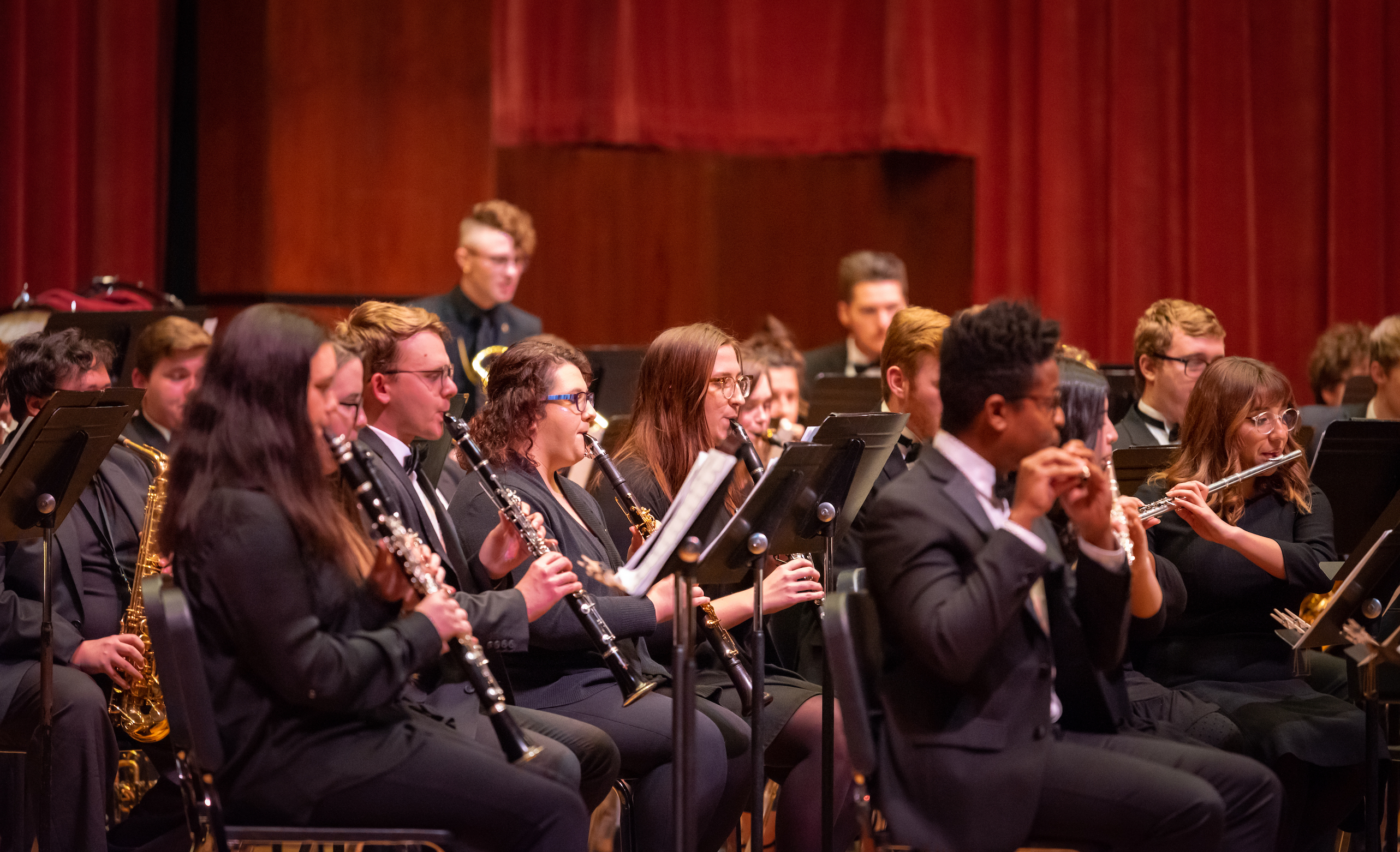 Side view of a wind band of students.  Far left is a saxophone, in the middle is a line of clarinets, and on the far right is a line of flutes.