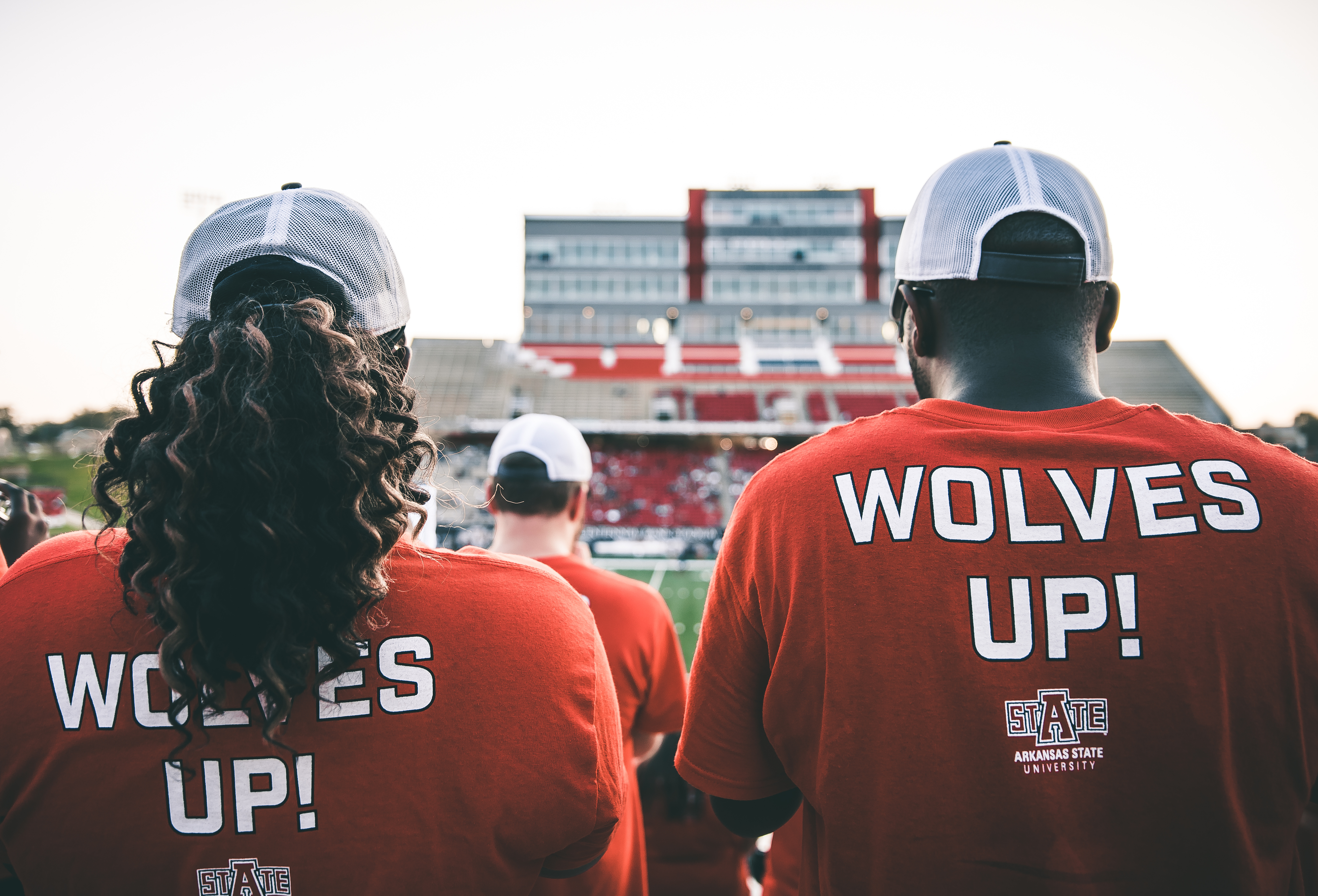 The back of two band students in t-shirts that say Wolves Up.  They are performing in Centennial Bank Stadium.