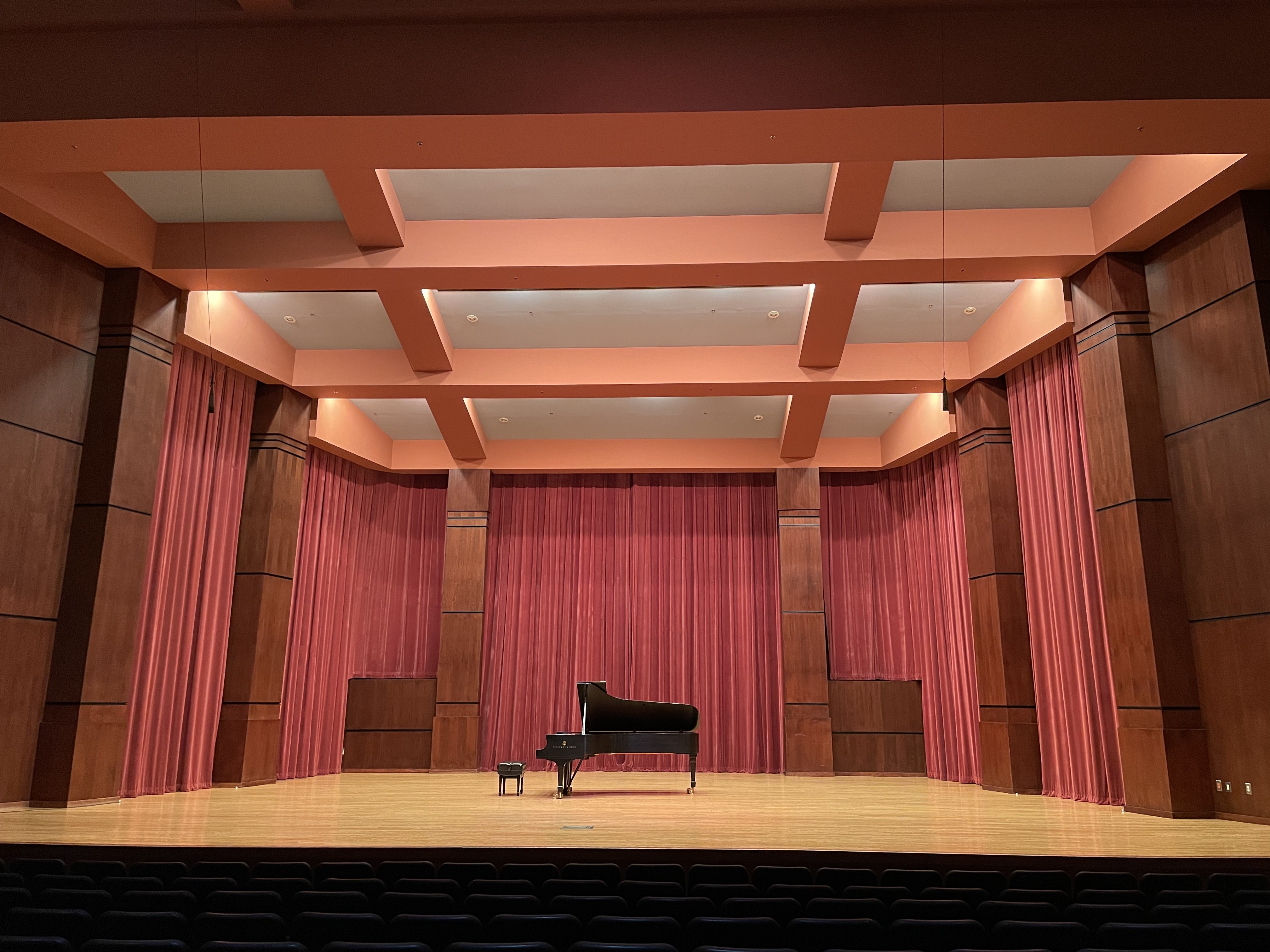 A view of the Riceland Hall stage from the audience.  There is an open grand piano on stage.  The celing is peach and white.  There are red curtains drawn broken up by wood grain columns.
