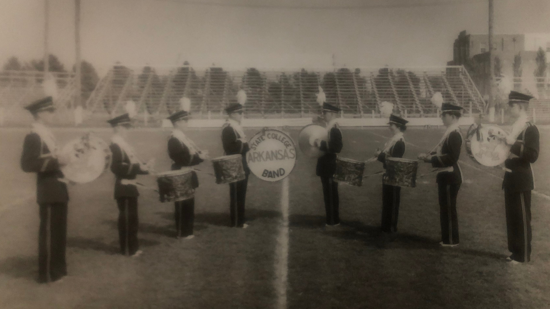 Historical photo of the marching band drumline on the field in a V shape.  The middle drum head says Arkansas State College Band.  There are four snare drums, a cymbol player, and two other bass drums.