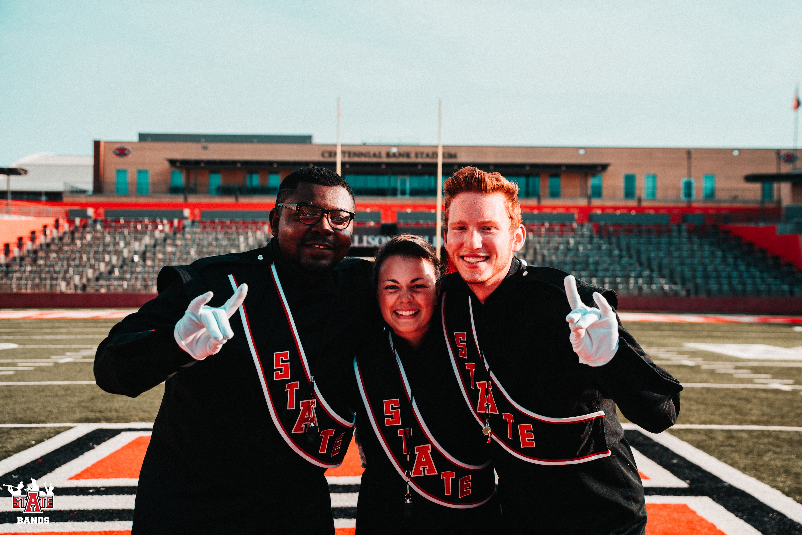 Three A-State drum majors in Centennial Bank Stadium giving a Wolves Up hand sign.