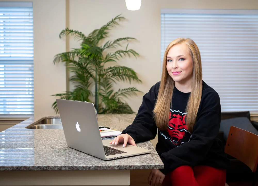 A-State student on laptop posing for a picture in a redwolf sweater