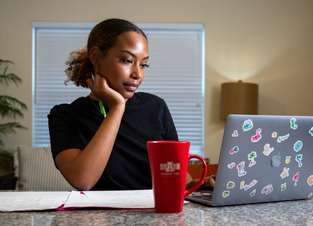 A-State student working on laptop with an A-State red coffee mug in front of her.
