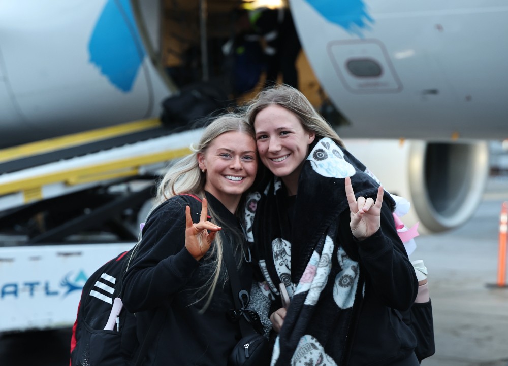 Two students giving wolves up hand gesture in front of plane being loaded with luggage