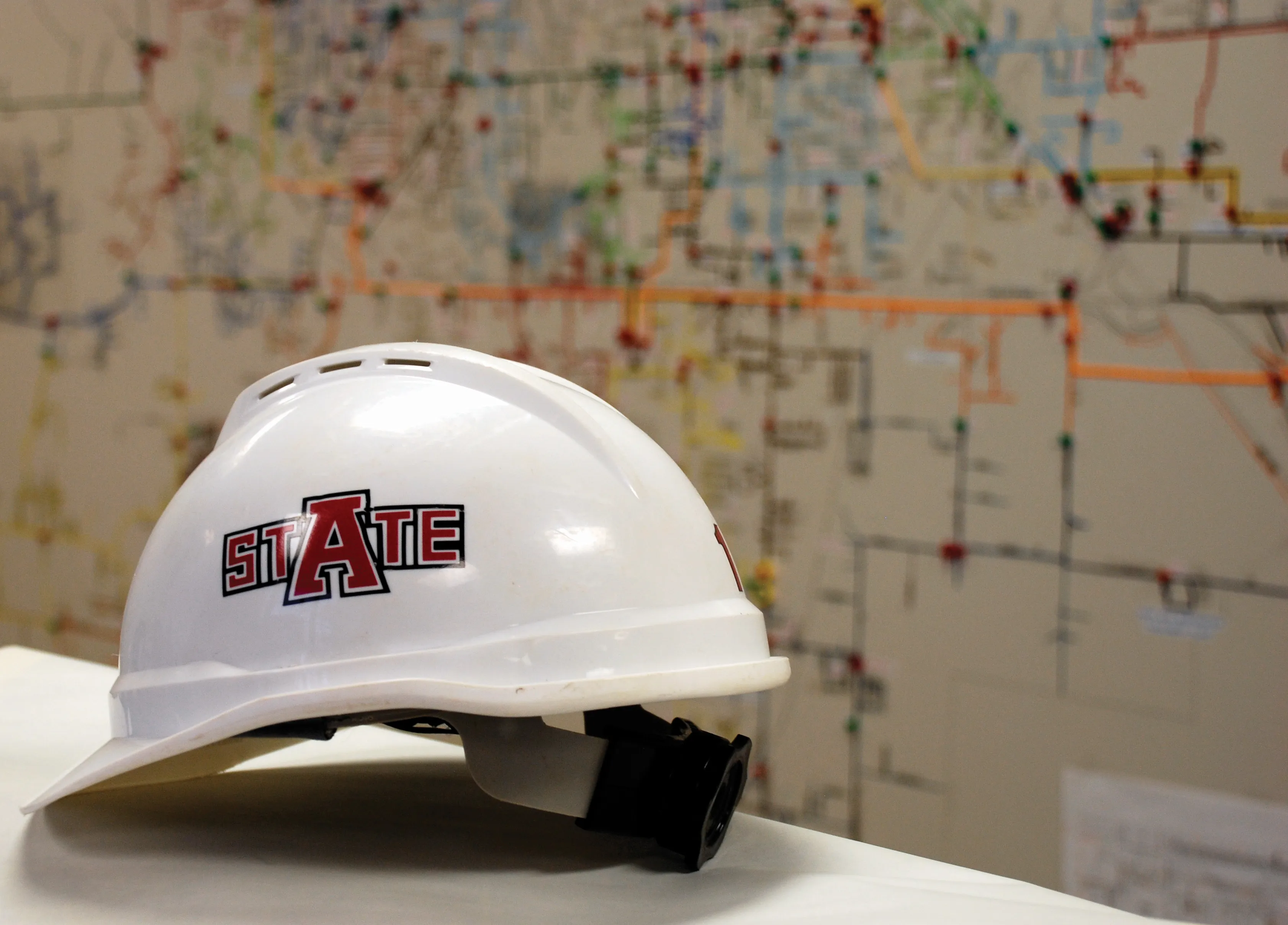 A-State logo on white hard hat with a blurry city infrastructure map background.