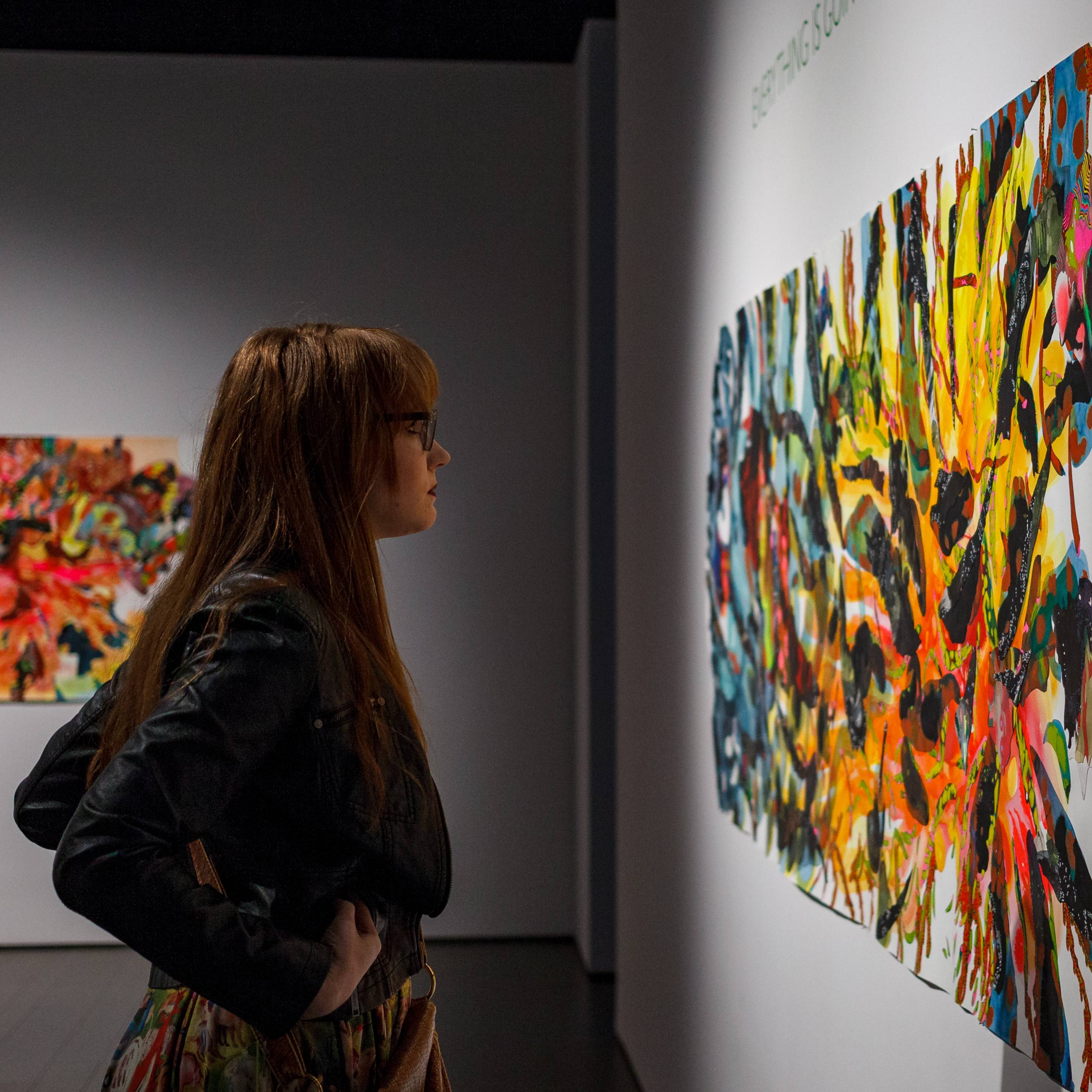 A person standing in an art gallery closely observing a large, colorful abstract painting on a white wall, with another vibrant artwork visible in the background.