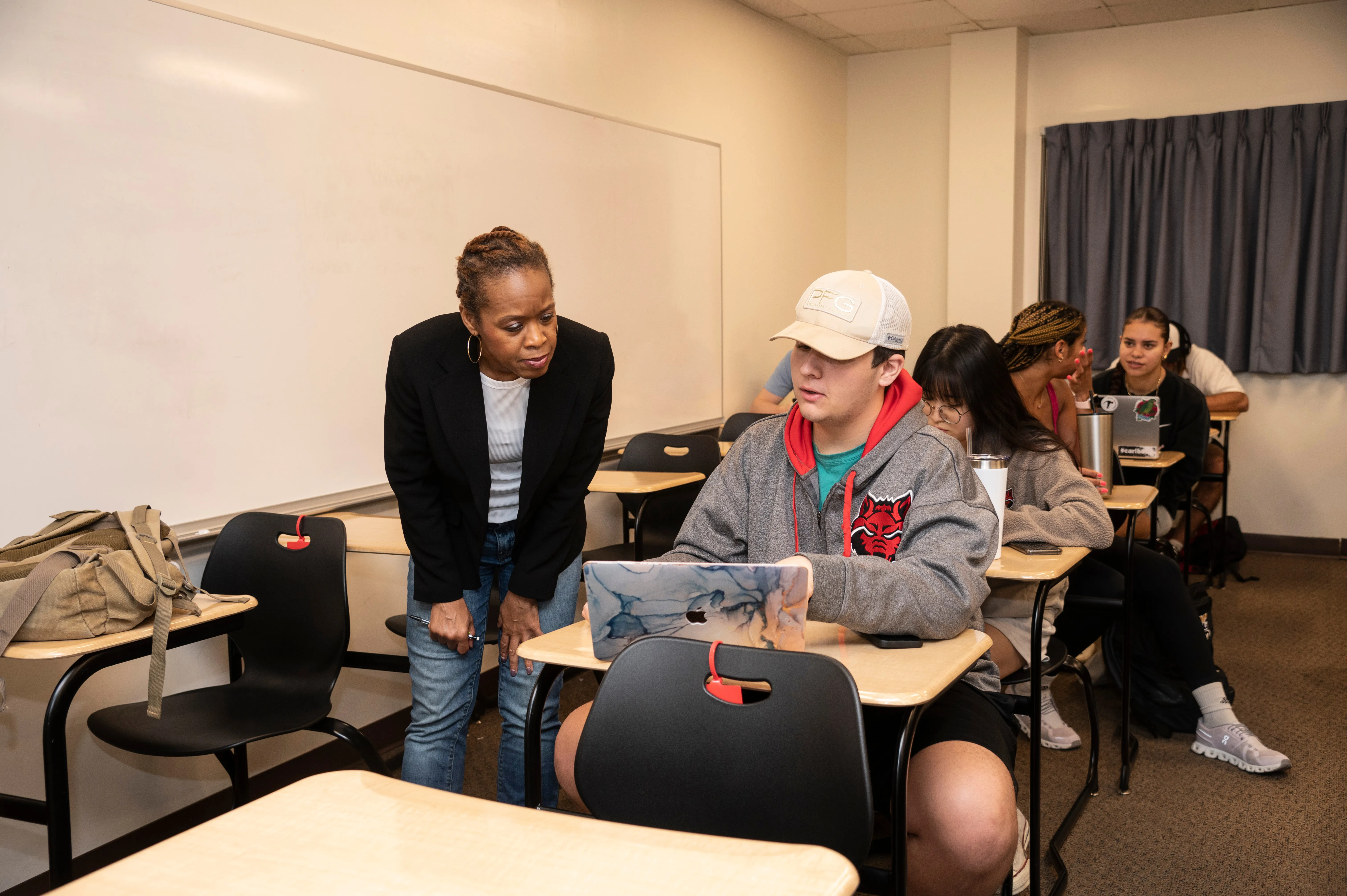In focus student in communication disorders class with redwolf sweater with other students in background and teacher standing over desk helping.