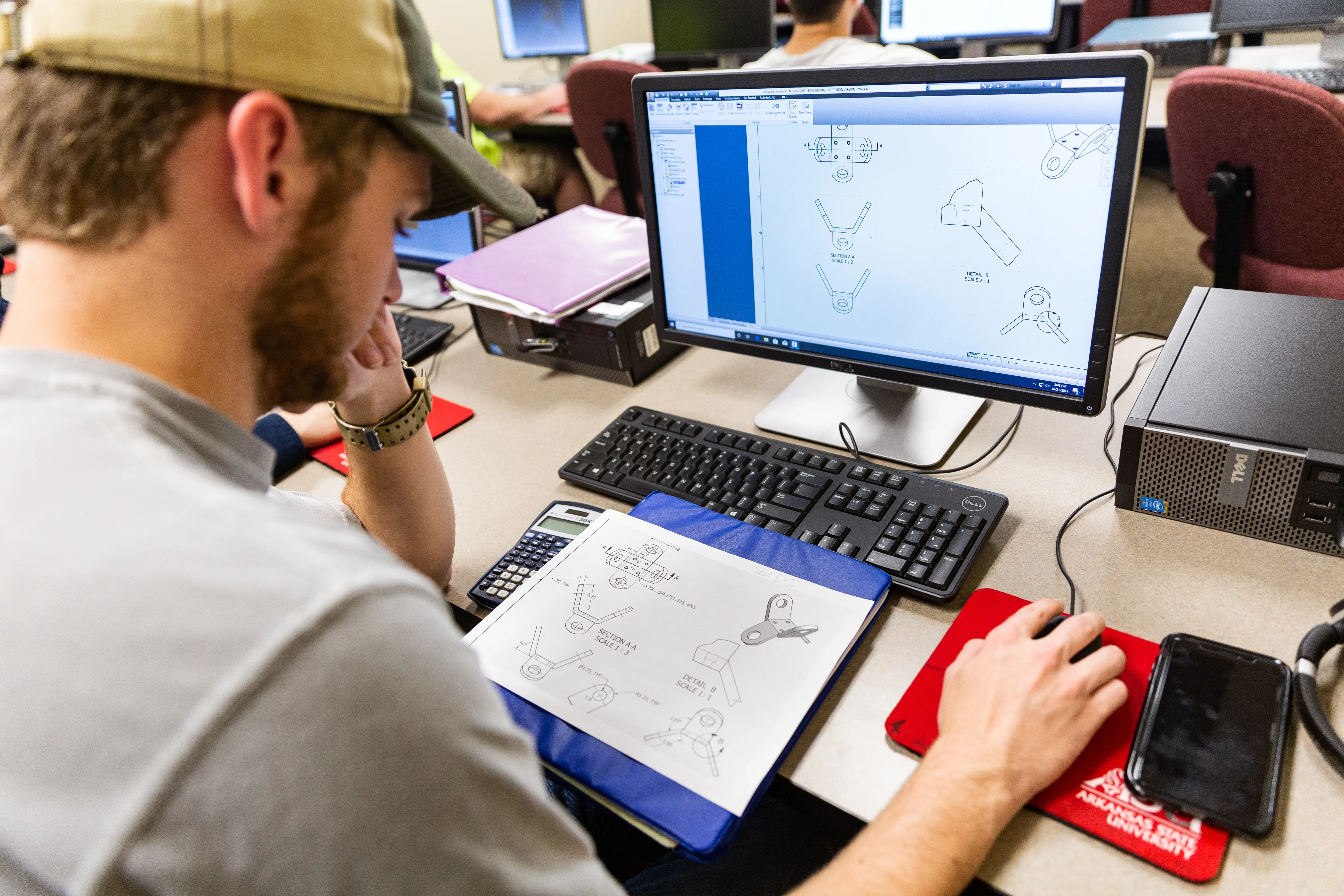 A-State student in Arkansas State shirt working on machine parts on a computer.