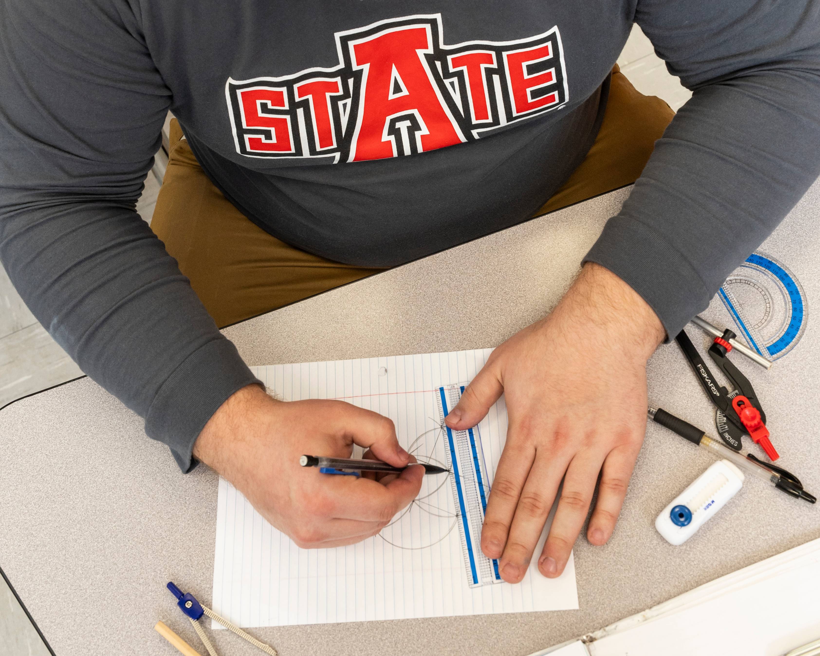 Student wearing an A-State shirt sketching on a piece of paper.