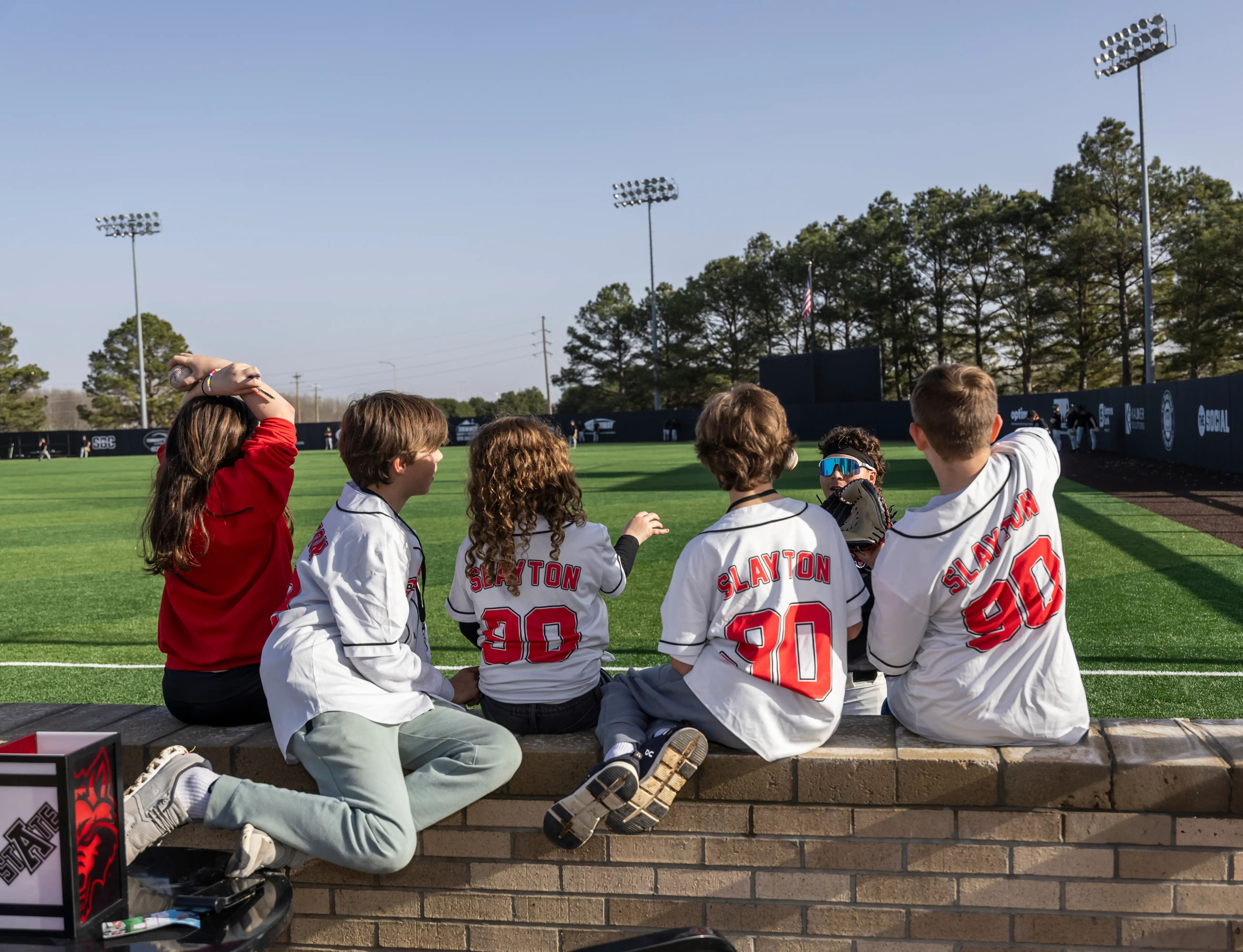 Five kids in jerseys sit on the edge of a wall at the A-State baseball field.