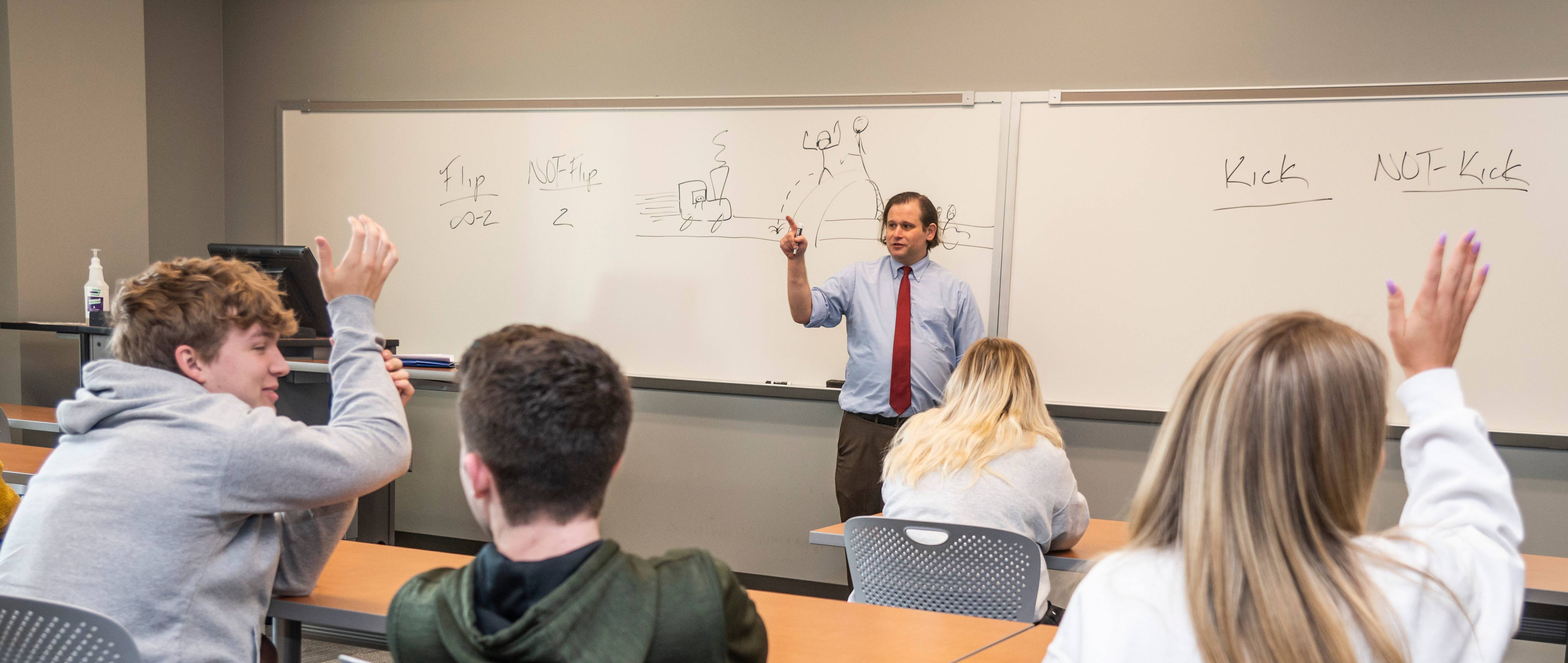 Professor in front of a whiteboard, facing the classroom and teaching. Students in the foreground are raising their hands.