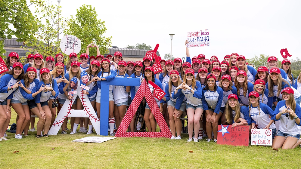 Large group wearing blue shirts and red hats posing with oversized Greek letters and signs outdoors during Bid Day.