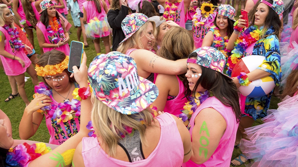 Group of people wearing bright pink shirts and floral accessories hugging and celebrating outdoors during Bid Day.