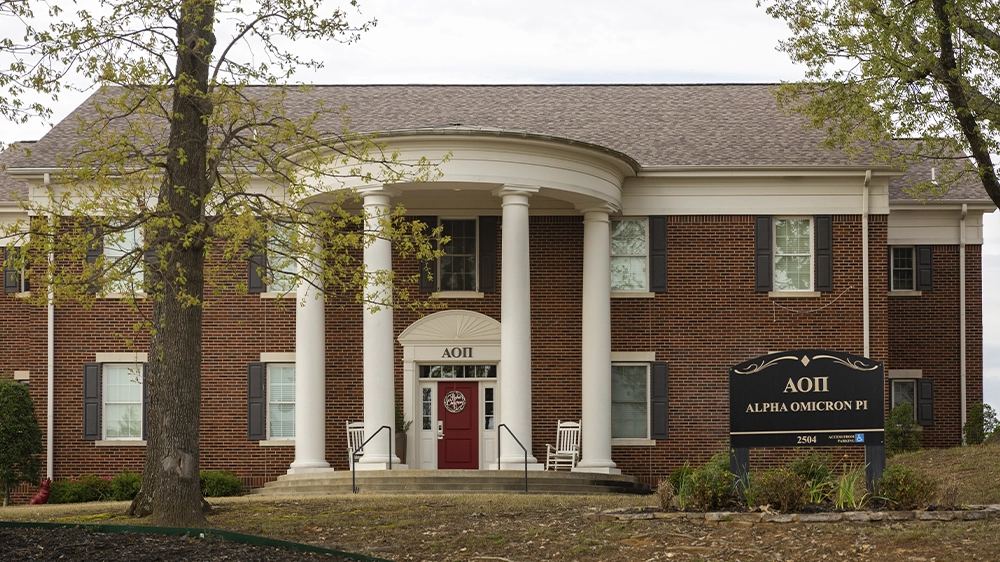 Two-story red brick sorority house with a curved portico supported by white columns. A black sign in front reads “ΑΟΠ Alpha Omicron Pi.” The front door is red, and the house is surrounded by trees and landscaping.