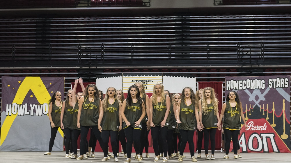Group of performers in matching dark tank tops and leggings standing in formation on an indoor stage with themed backdrops.