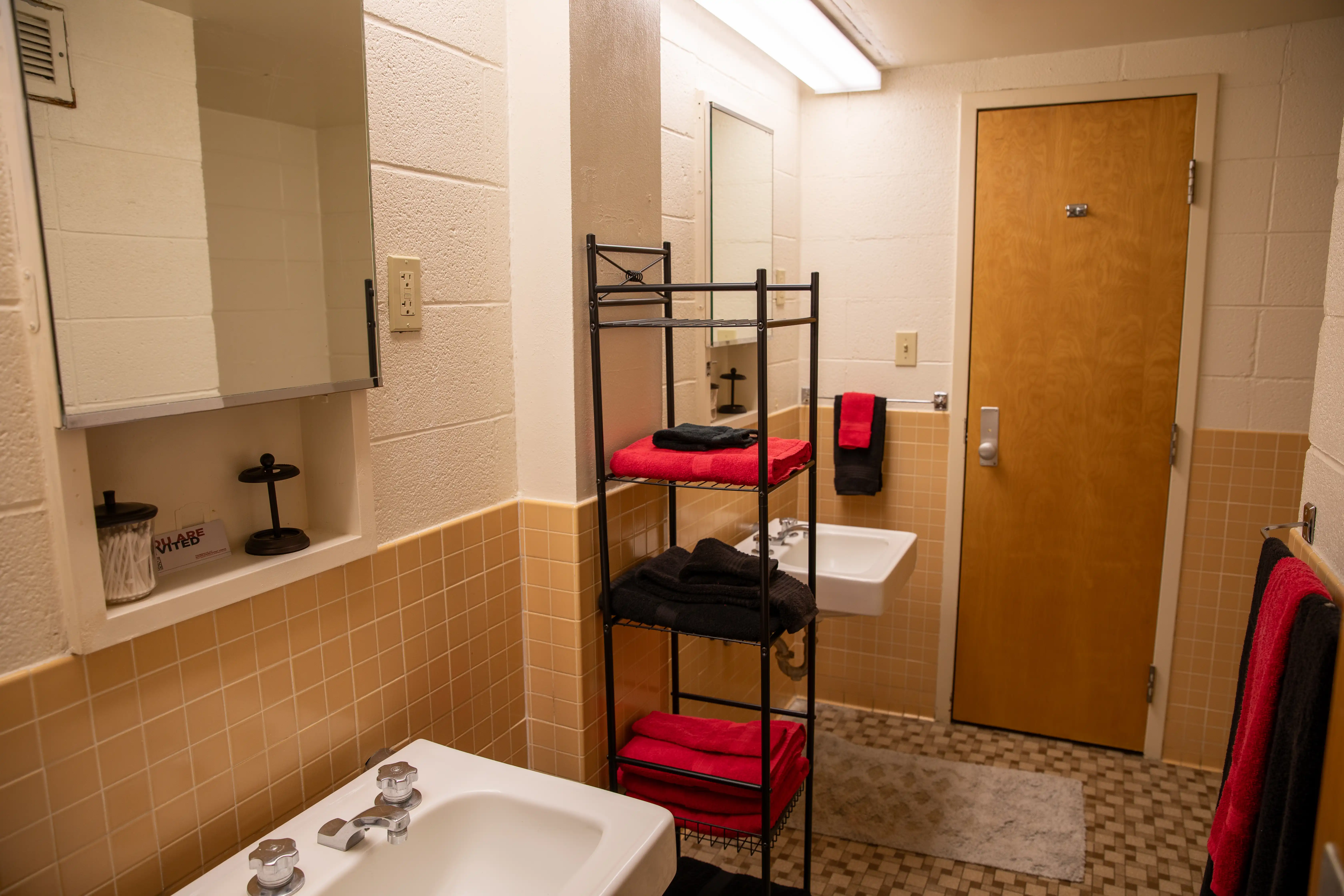 A bathroom with two sinks, two mirros, a shelving unit and tan-colored tile.