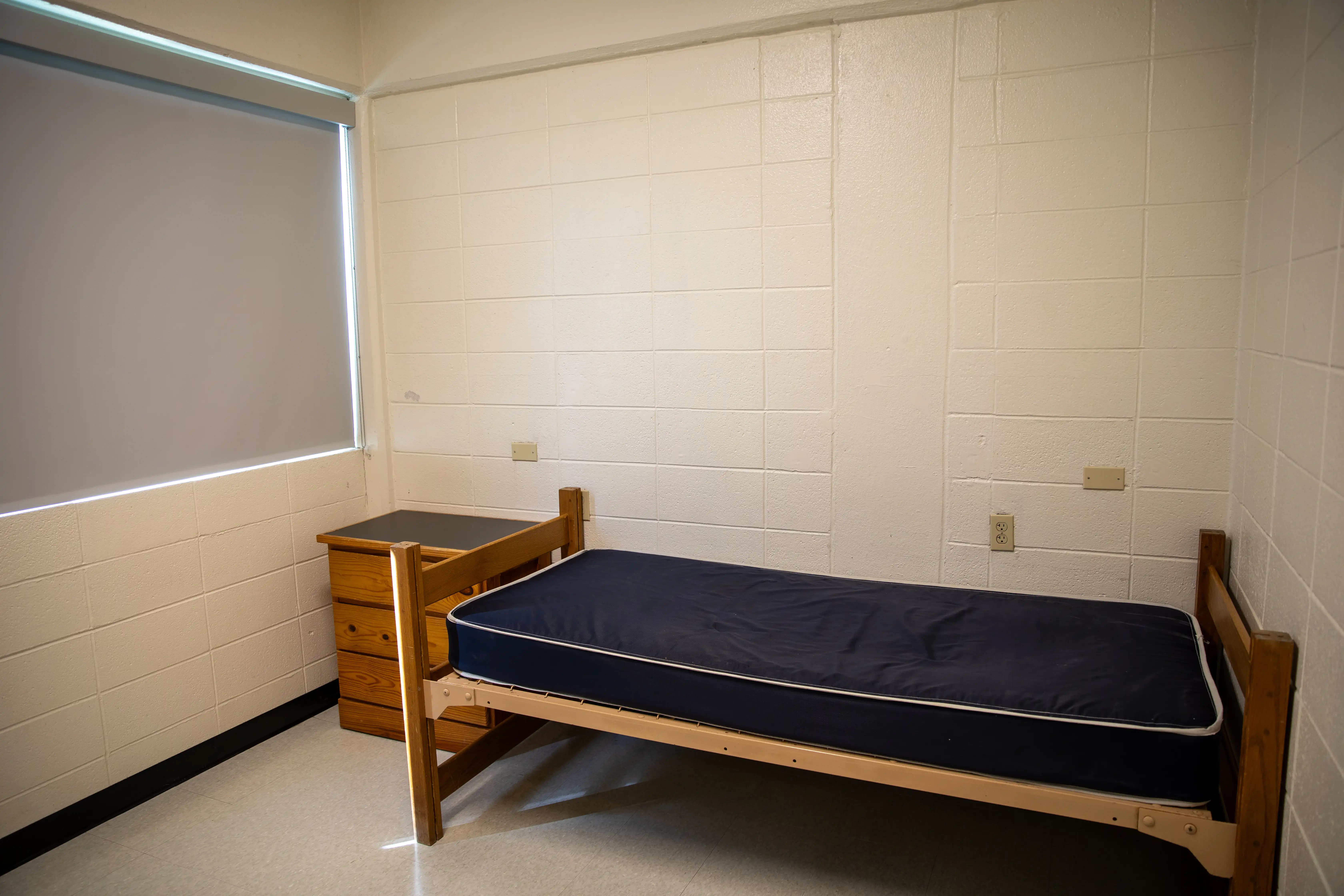 Twin-size bed frame with mattress and small chest of drawers in empty room.