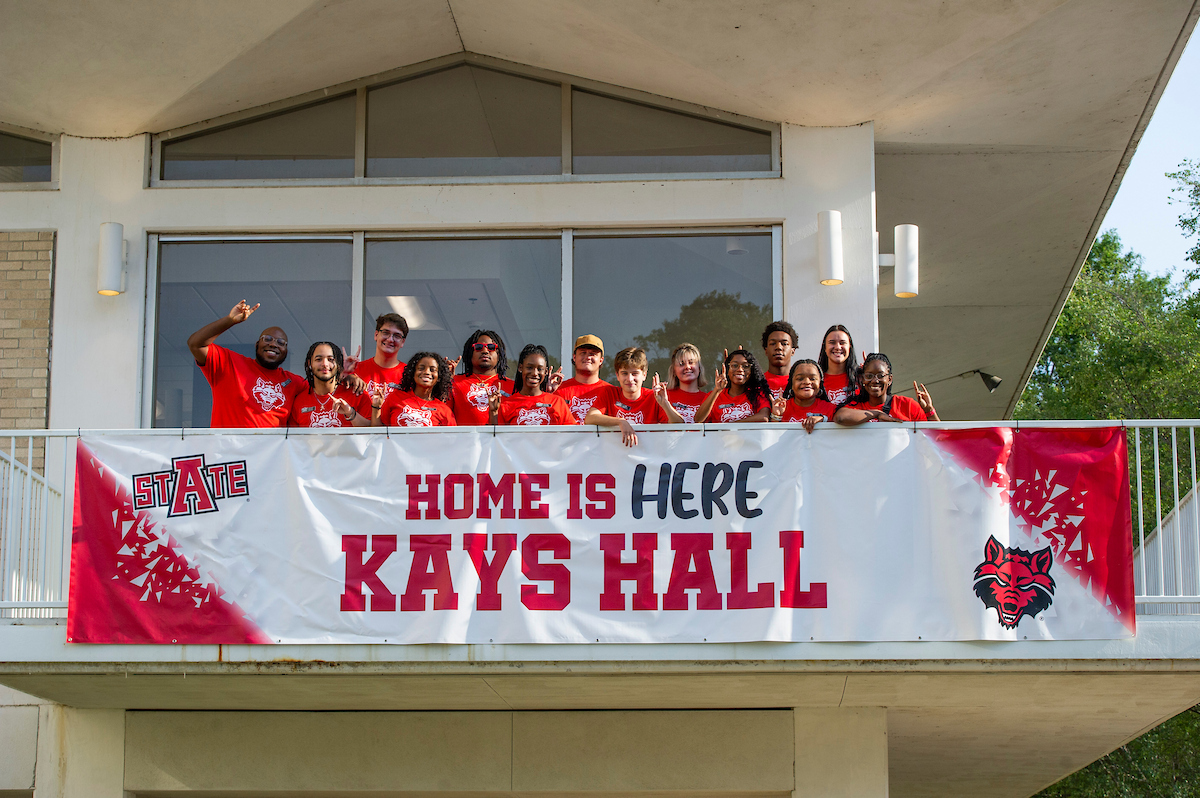 Students in red shirts pose on Kays Hall balcony behind a “Home is Here” banner at A-State.