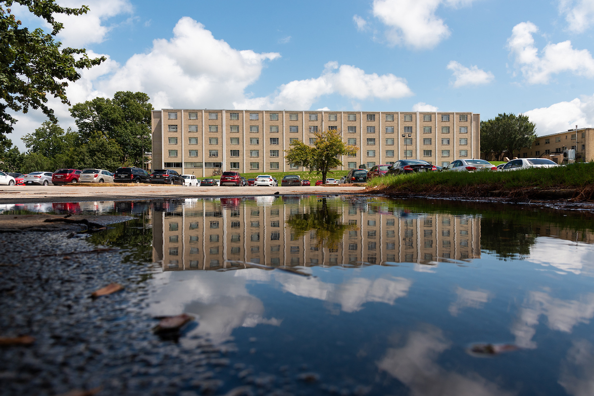 A-State’s University Hall and parked cars reflected in a rain puddle under a blue sky with clouds.