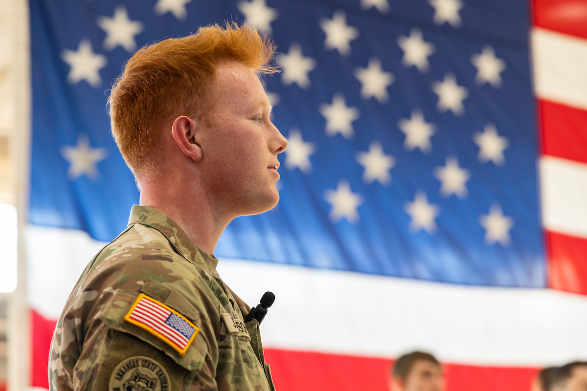 A-State ROTC student in uniform stands in front of large American flag during ceremony.