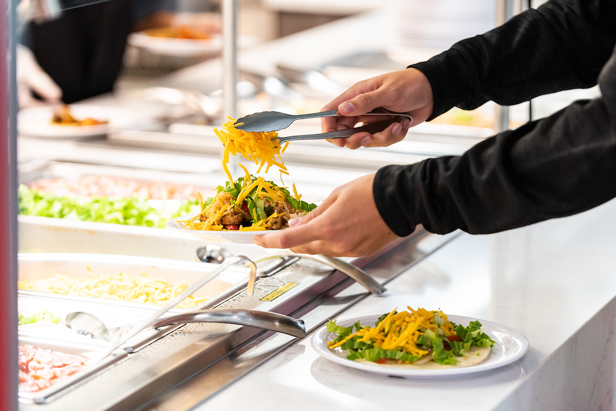 A-State student adds shredded cheese to tacos at a self-serve dining station on campus with fresh toppings available.