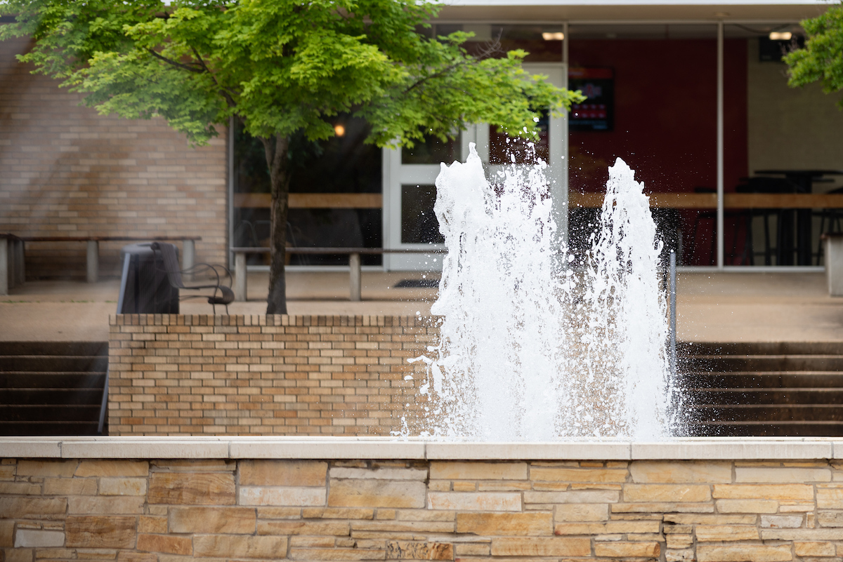 Water fountain splashes upward in front of a brick building on the A-State campus.
