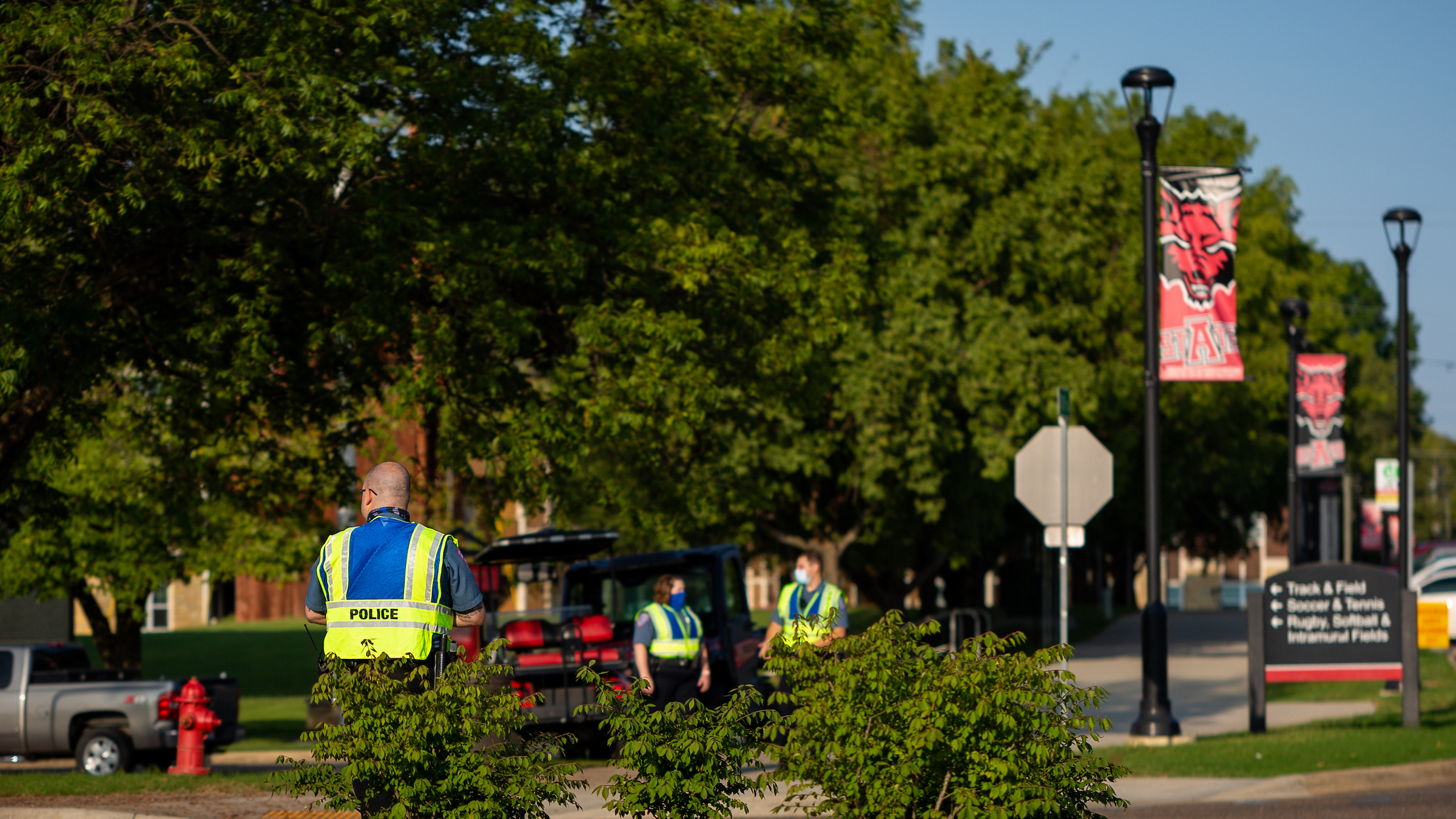 A-State University police officers patrol near the campus entrance under sunny skies and green trees.