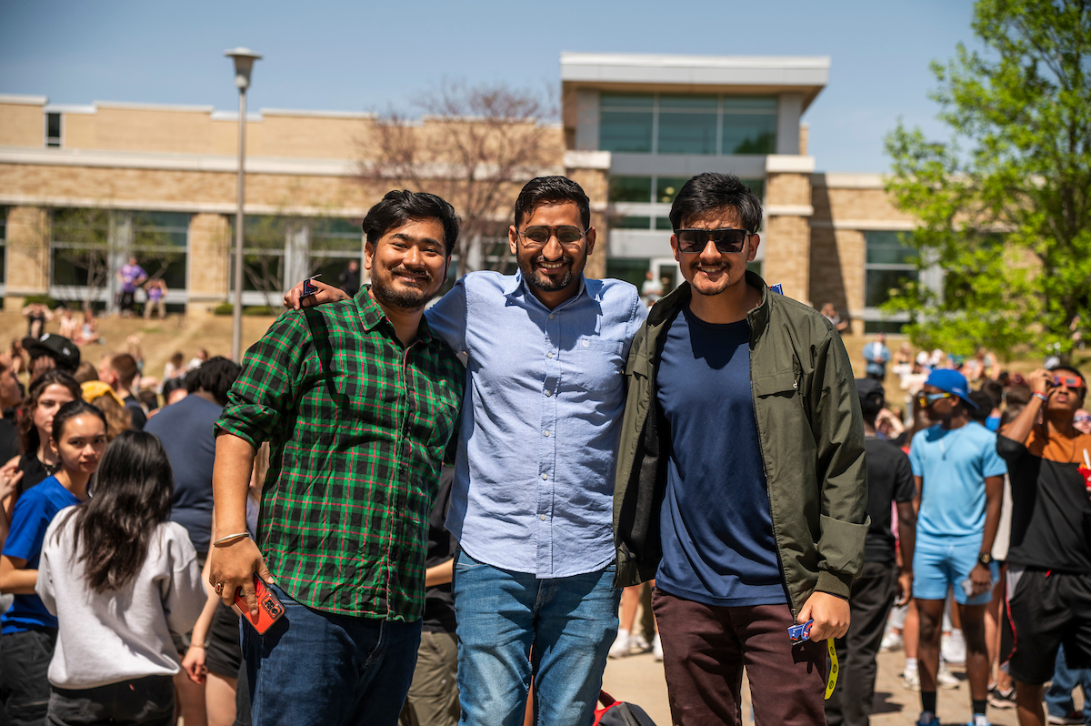 Three A-State students pose and smile outside during a sunny campus event.