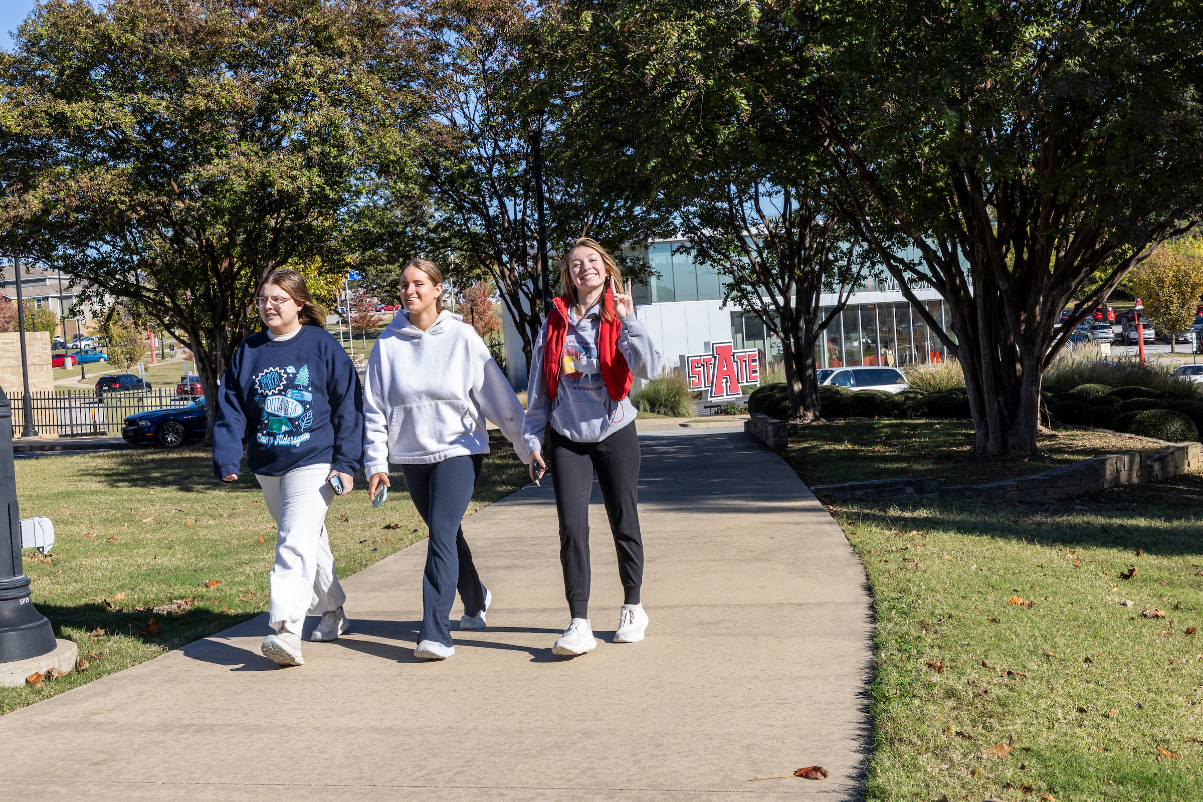 Students walking along a tree-lined campus pathway on a sunny fall day, with green grass and the Red Wolves Center building visible in the background.