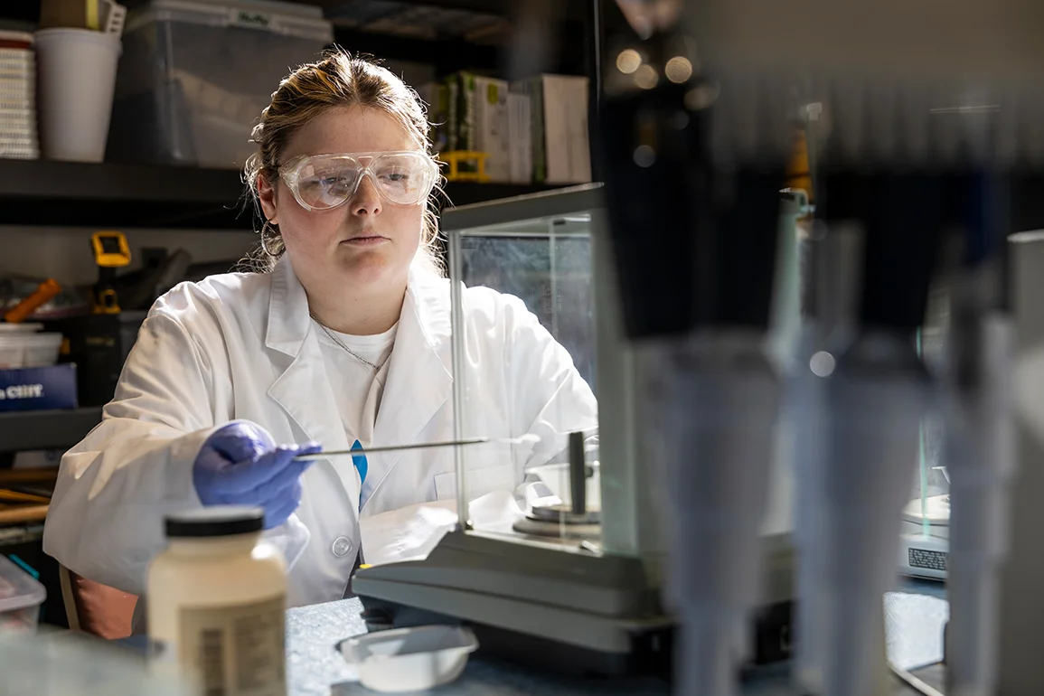 An Arkansas State University student wearing safety goggles and a lab coat conducts an experiment in a campus chemistry lab, carefully measuring a sample on a precision scale surrounded by scientific equipment.