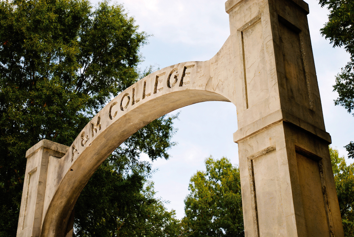 Historic A&M College arch at A-State surrounded by trees under a partly cloudy sky.