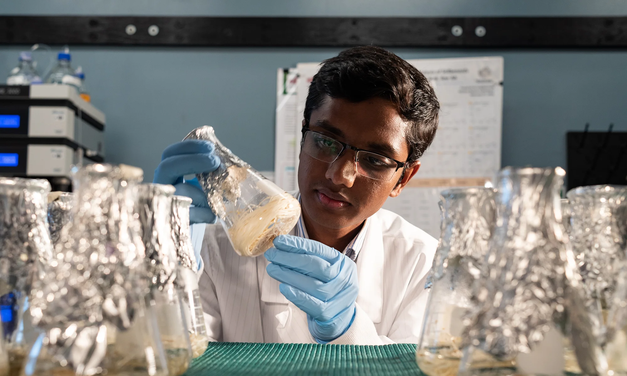 A student in a lab coat inspecting a beaker covered in tinfoil.