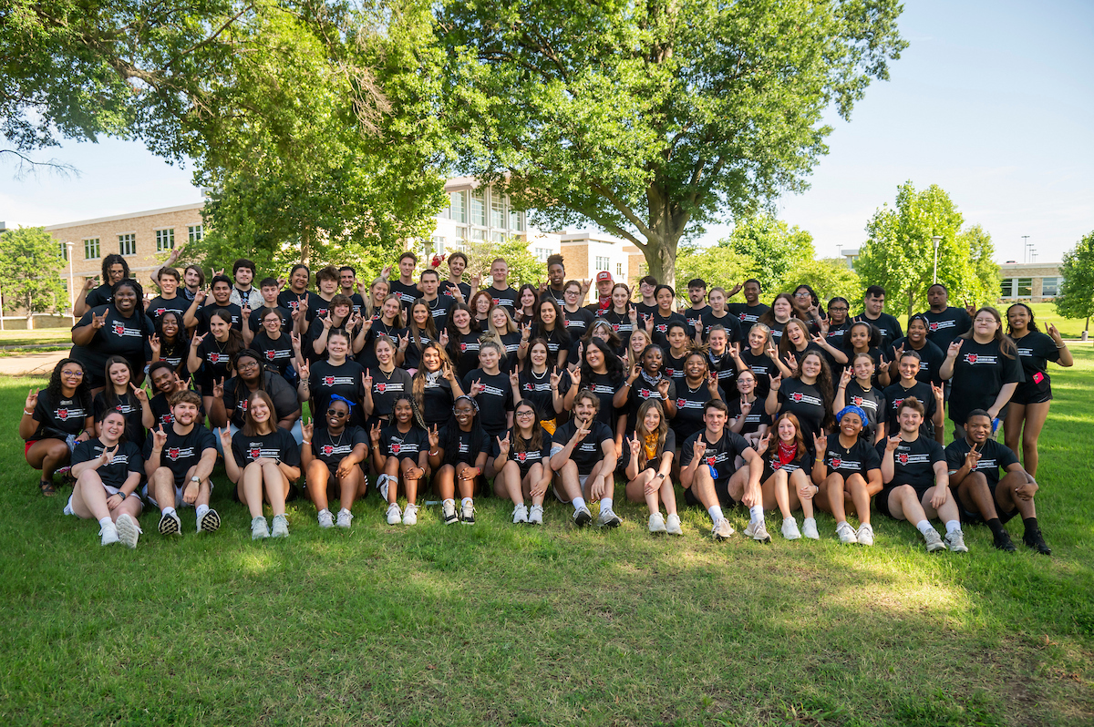 Large group of A-State orientation leaders posing together on campus lawn in matching shirts, smiling and throwing Red Wolves hand signs.
