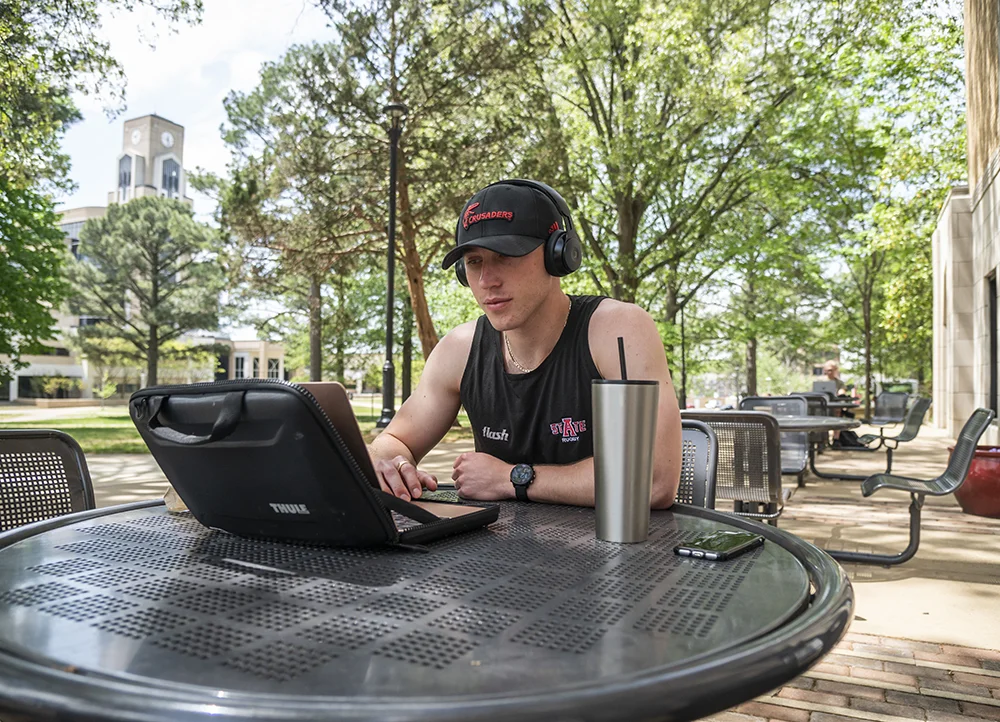 An A-State Rugby player using a laptop outside on campus.
