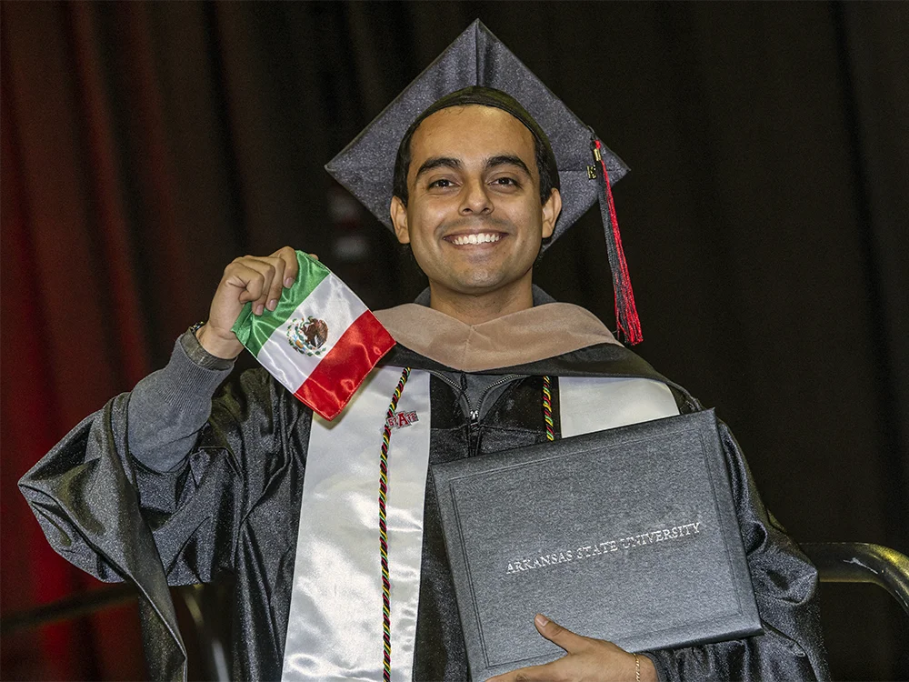 An A-State graduate holding his diploma and the flag of Mexico.