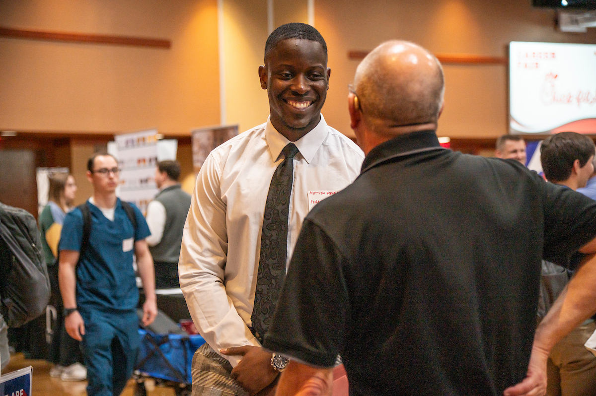 A professionally dressed student smiles while speaking with an employer at an indoor career fair.