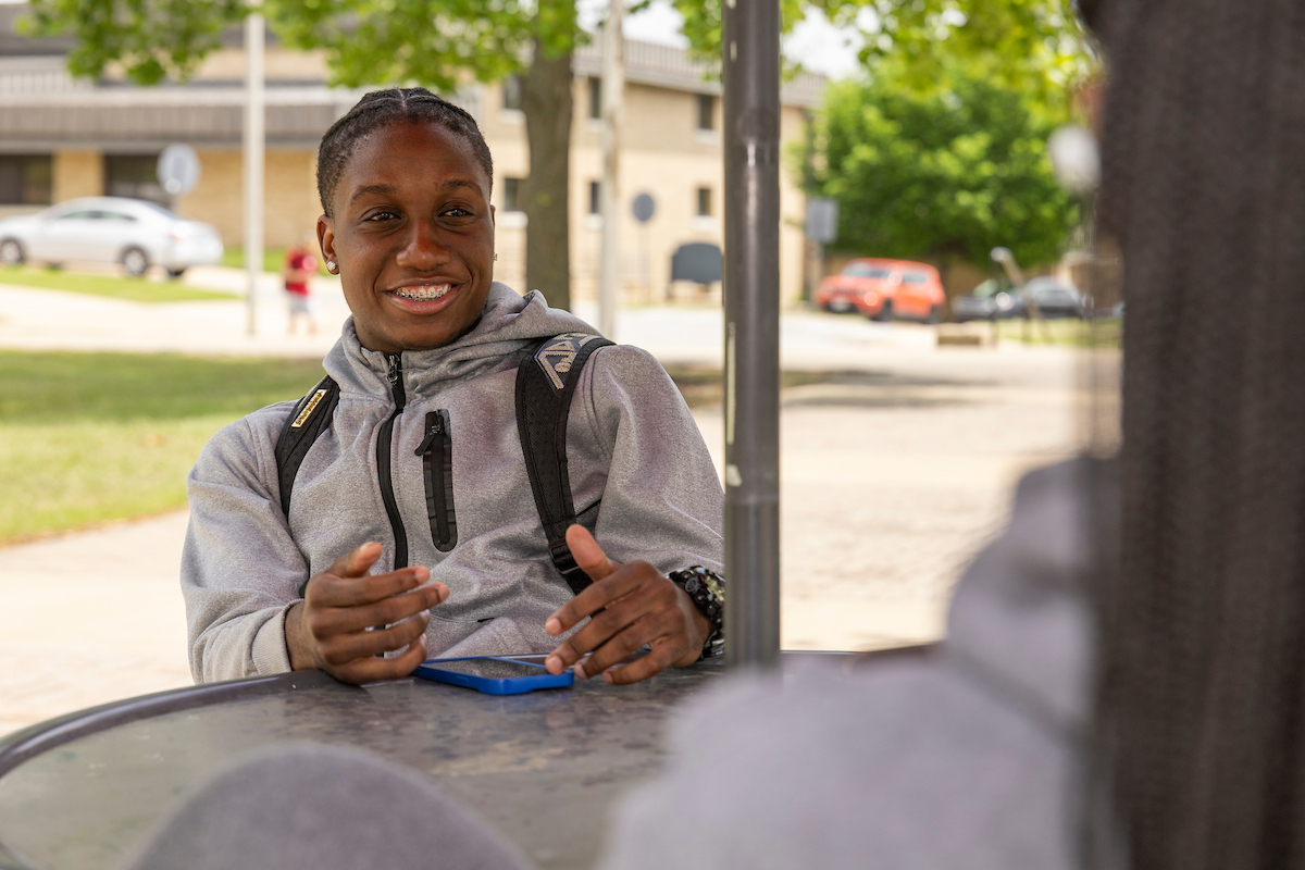 A student sitting at a table and talking to someone offscreen. 