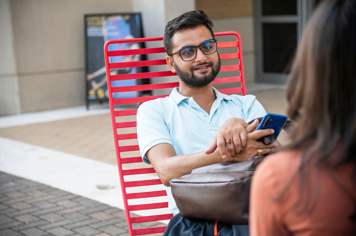 A-State student relaxes in a red chair during an outdoor chat, holding a phone and smiling.