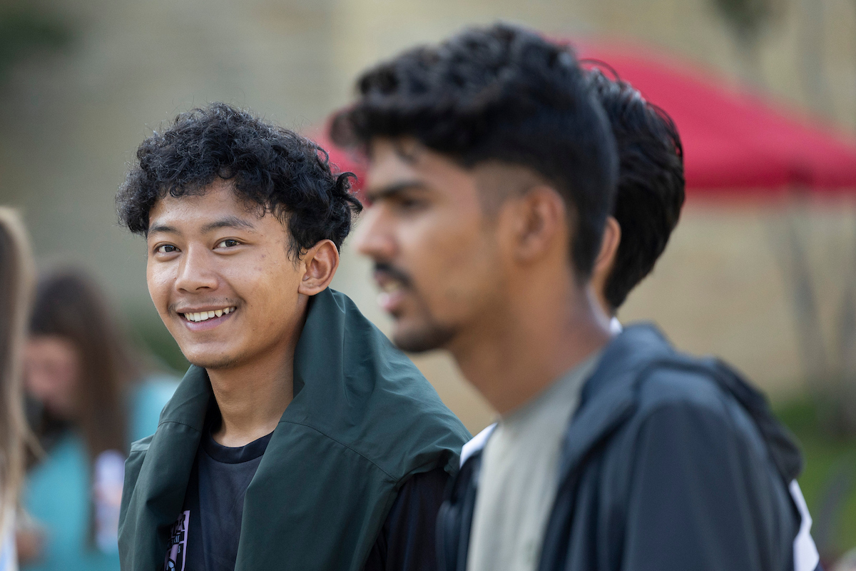 A smiling A-State student looks at the camera during a campus gathering, with other students chatting in the background.