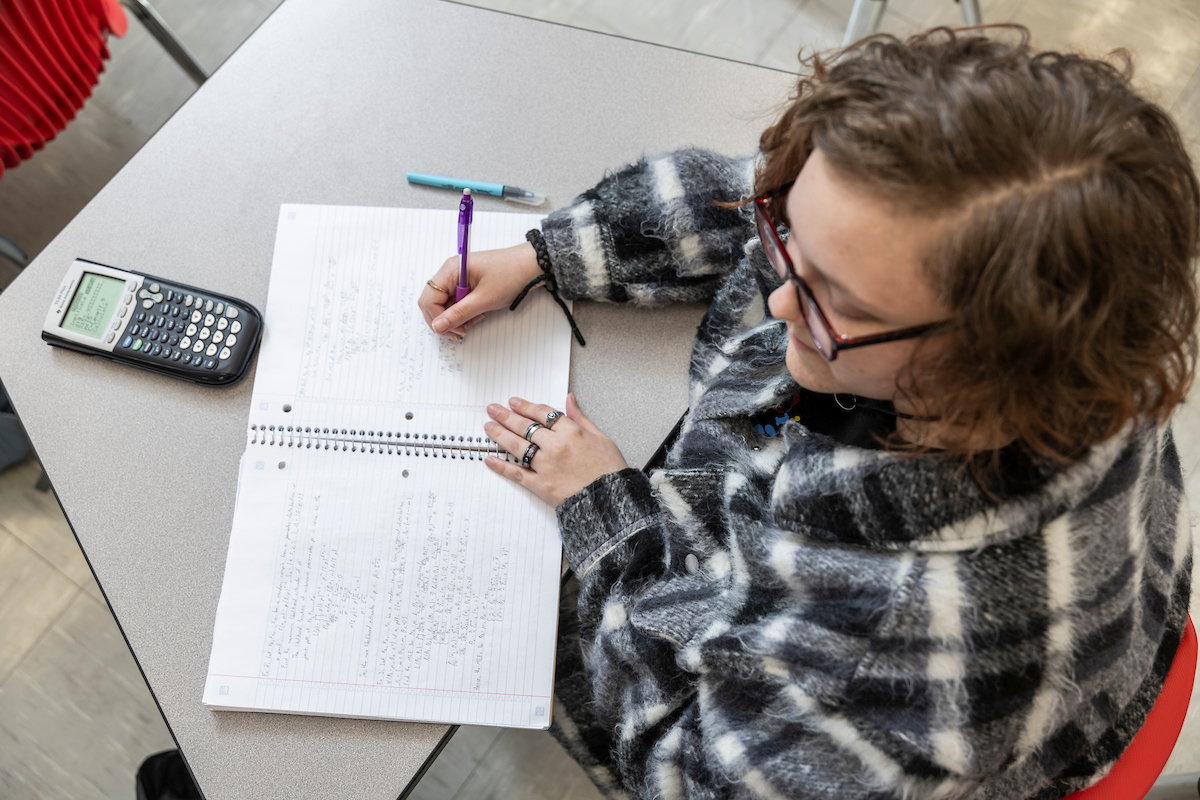 A-State student writing math problems in a notebook with a calculator on the table.