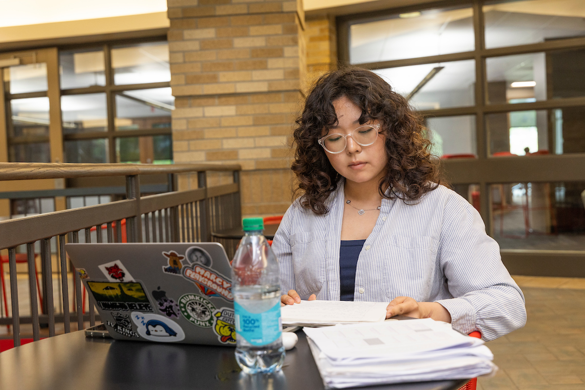 A-State student studies indoors with notes and a laptop, focused on reading while seated at a table with a water bottle and papers nearby.