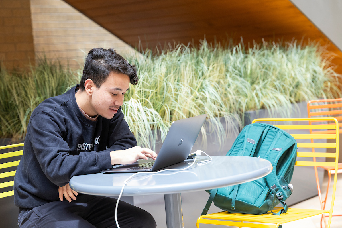 Student sits at a table in HHS atrium working on a laptop with a backpack on a nearby chair.