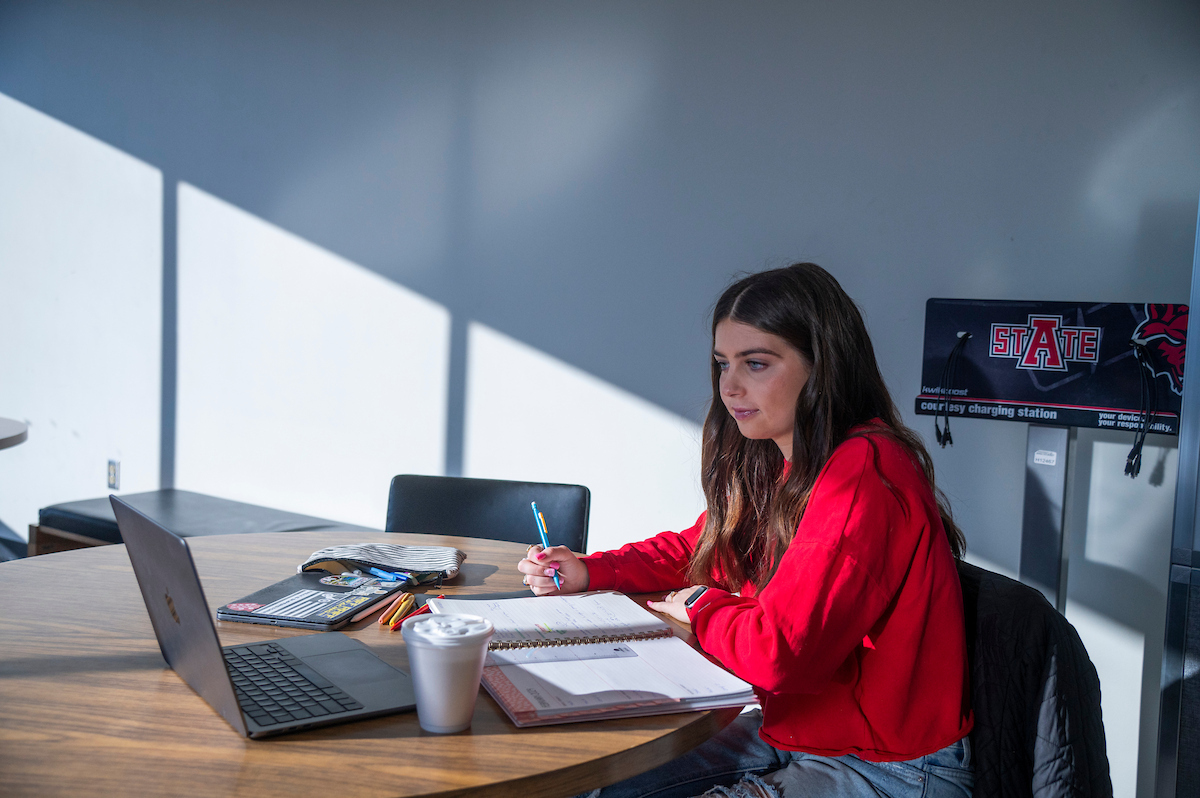 A student studies with a laptop and planner at a table near a charging station in a naturally lit campus space.