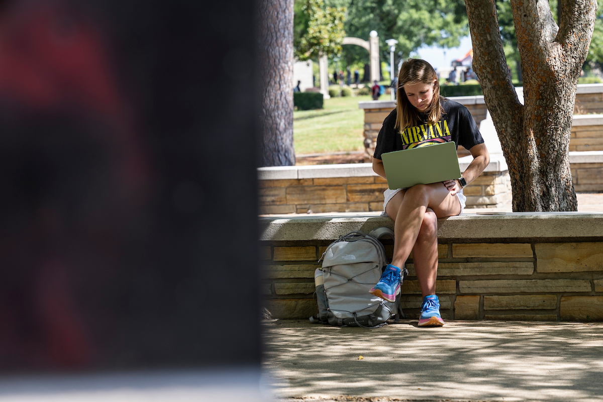 An student studies outside on a stone bench with a laptop and backpack, enjoying a shaded spot on campus.